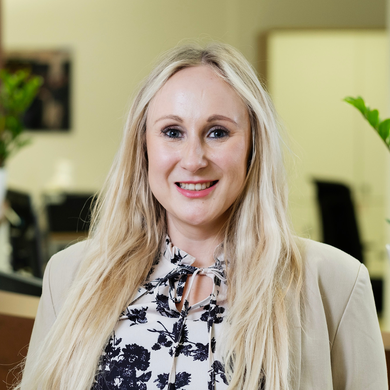 Eine Frau mit langen blonden Haaren lächelt in die Kamera. Sie trägt eine beige Jacke und eine helle Bluse mit schwarzem Blumenmuster. Im Hintergrund ist ein unscharfes Büro mit Pflanzen zu sehen. Eine Frau mit langen blonden Haaren lächelt in die Kamera. Sie trägt eine beige Jacke und eine helle Bluse mit schwarzem Blumenmuster. Im Hintergrund ist ein unscharfes Büro mit Pflanzen zu sehen.