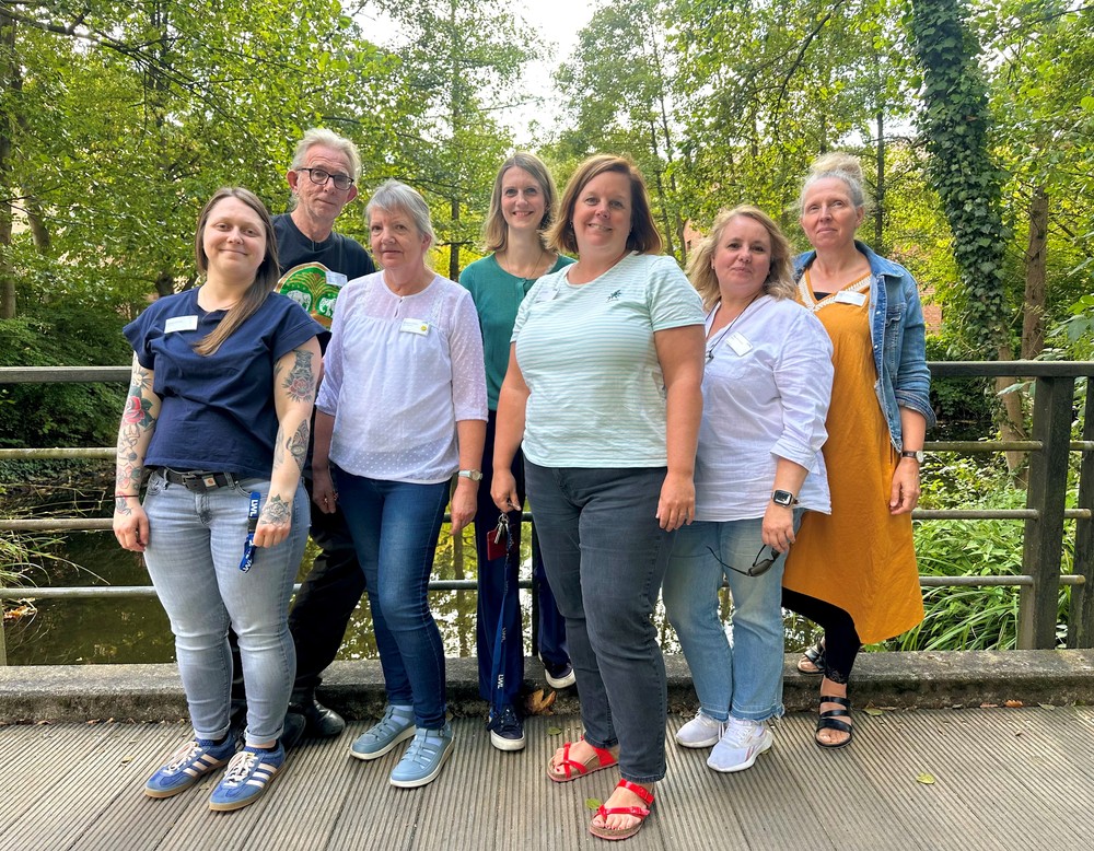 Eine Gruppe von sieben Personen steht im Freien auf einer Brücke und blickt in die Kamera. Im Hintergrund sind Bäume und ein Bach zu sehen. Eine Gruppe von sieben Personen steht im Freien auf einer Brücke und blickt in die Kamera. Im Hintergrund sind Bäume und ein Bach zu sehen.