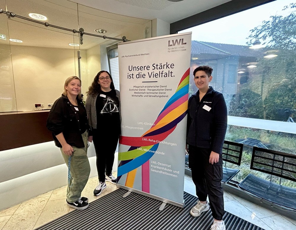 Drei Personen stehen in einem Eingangsbereich neben einem Roll-up mit der Aufschrift „Unsere Stärke ist die Vielfalt“. Das Plakat zeigt farbige Streifen und Logos des LWL. Im Hintergrund sind Glaswände, Stühle und ein Blick nach draußen zu sehen. Drei Personen stehen in einem Eingangsbereich neben einem Roll-up mit der Aufschrift „Unsere Stärke ist die Vielfalt“. Das Plakat zeigt farbige Streifen und Logos des LWL. Im Hintergrund sind Glaswände, Stühle und ein Blick nach draußen zu sehen.