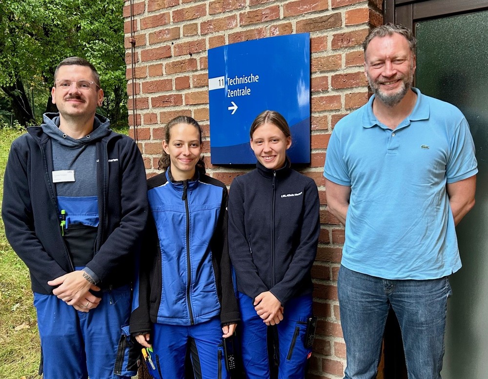 Vier Personen stehen lächelnd vor einem Backsteingebäude mit einem Schild „Technische Zentrale“. Zwei von ihnen tragen blaue Arbeitskleidung, die anderen Freizeitkleidung. Vier Personen stehen lächelnd vor einem Backsteingebäude mit einem Schild „Technische Zentrale“. Zwei von ihnen tragen blaue Arbeitskleidung, die anderen Freizeitkleidung.