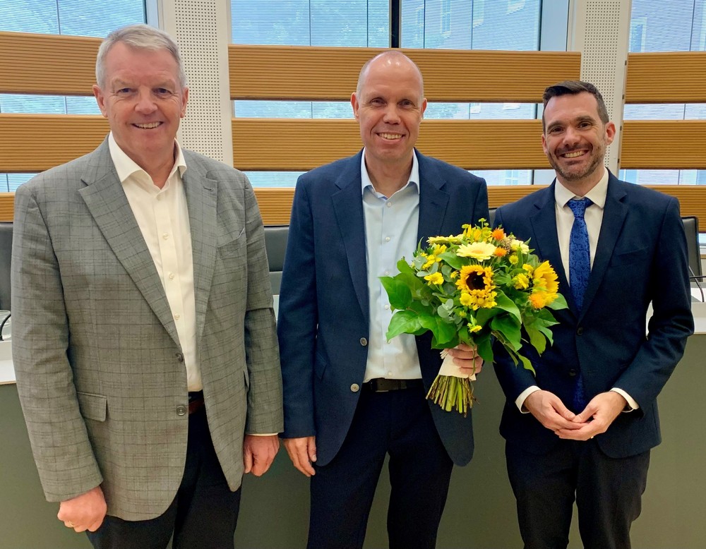 Drei Männer in Anzügen stehen nebeneinander und lächeln in die Kamera. Der Mann in der Mitte hält einen Strauß gelber Blumen mit Sonnenblumen in der Hand. Drei Männer in Anzügen stehen nebeneinander und lächeln in die Kamera. Der Mann in der Mitte hält einen Strauß gelber Blumen mit Sonnenblumen in der Hand.