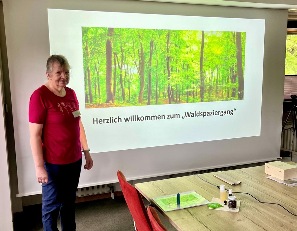 Eine Frau steht neben einer Leinwand, auf der eine Waldszene mit dem Text "Herzlich willkommen zum Waldspaziergang" projiziert wird. Eine Frau steht neben einer Leinwand, auf der eine Waldszene mit dem Text "Herzlich willkommen zum Waldspaziergang" projiziert wird.